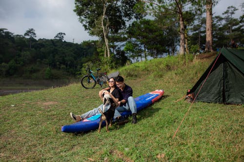 Photo of a Couple with a Dog Sitting on a Kayak next to a Tent