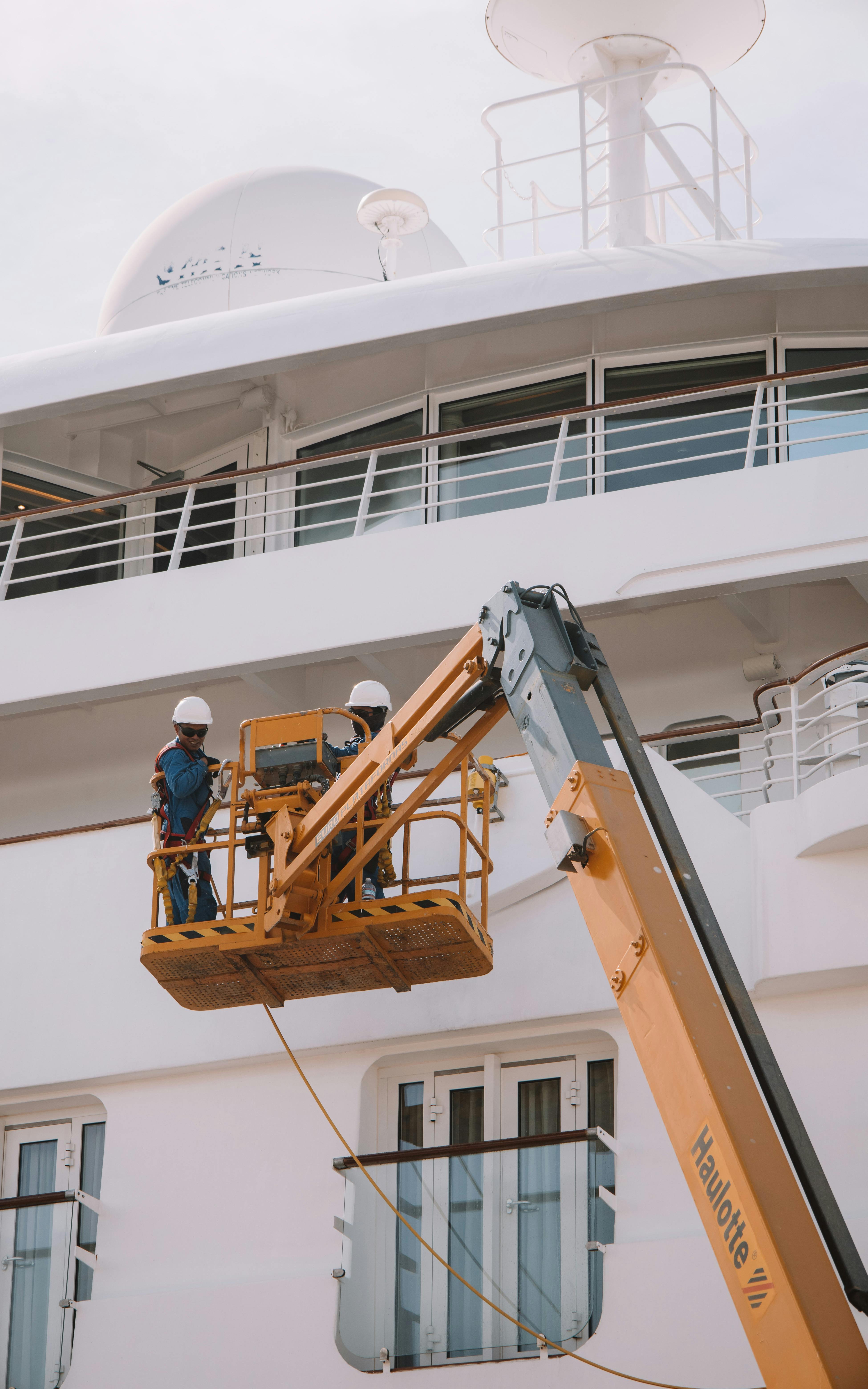 Two workers in a yellow lift platform repairing a white yacht.