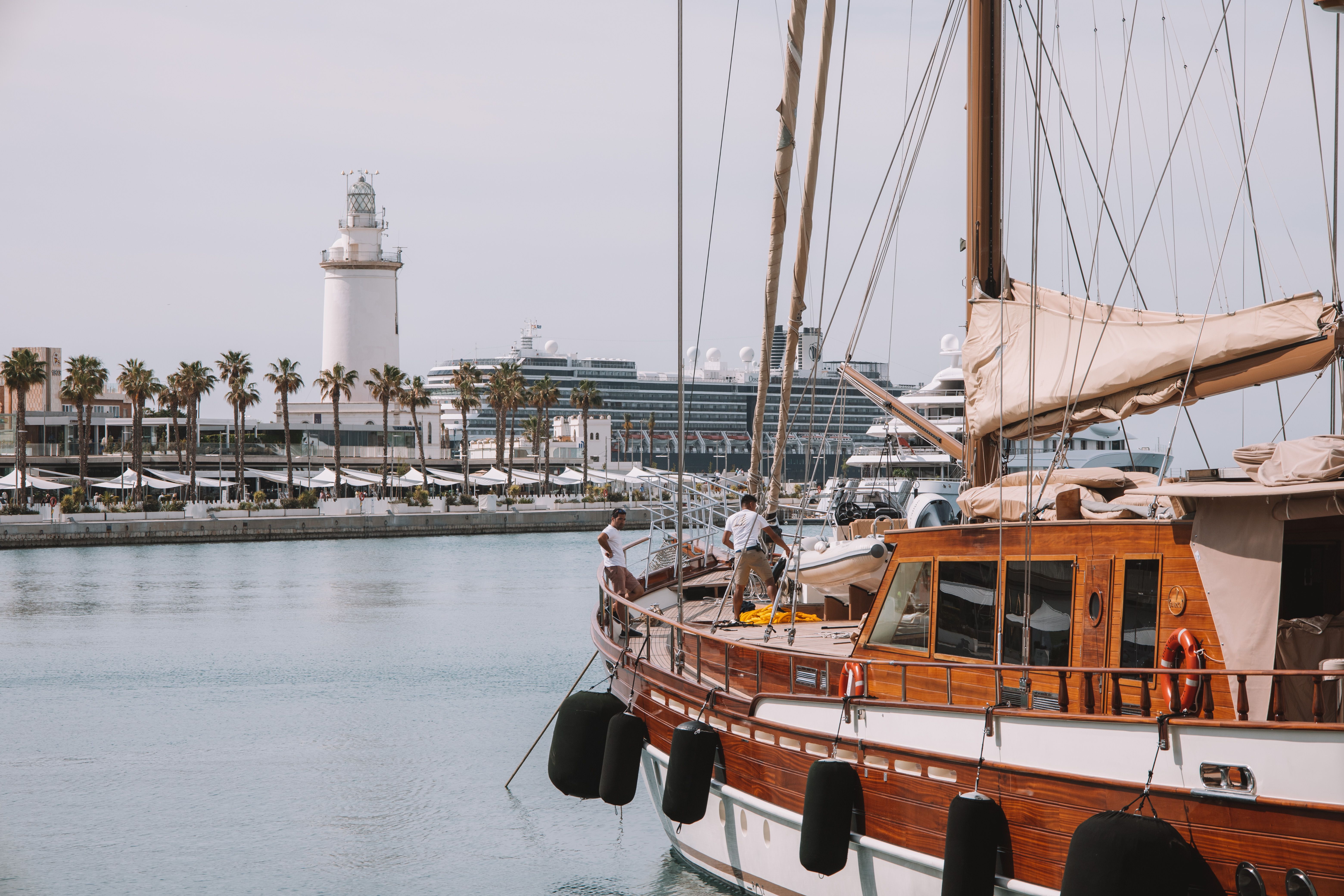 A wooden sailboat moored at Malaga port with the lighthouse and quayside terraces behind