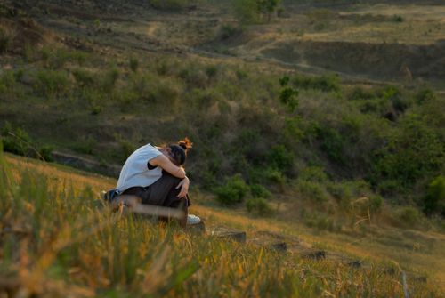 Free A woman sits alone on a grassy hill during a peaceful summer sunset. Stock Photo