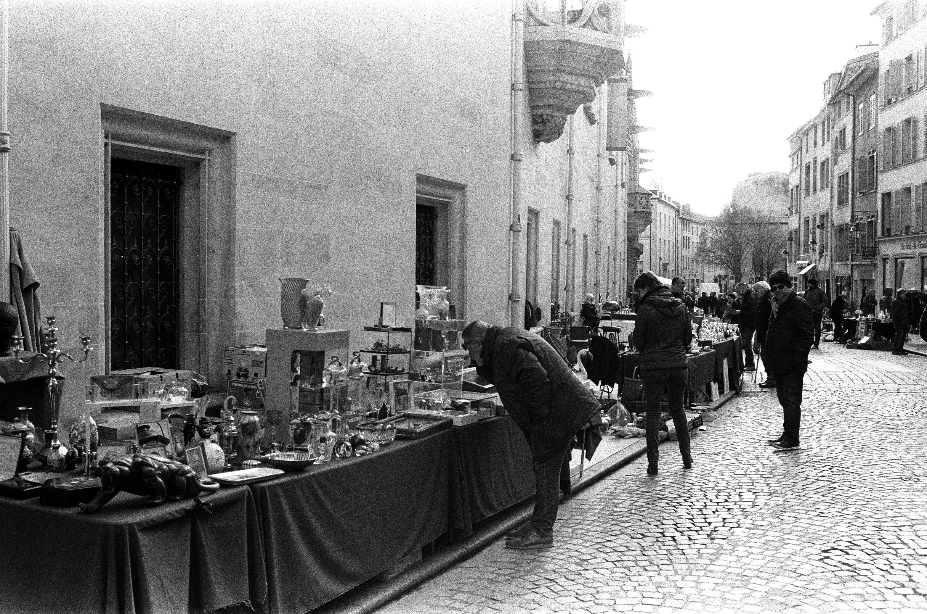 A black and white photo of a sidewalk market with tables of antiques and vintage objects, and shoppers browsing.