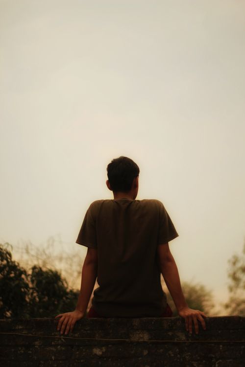 Free A man in a t-shirt sits on a wall, gazing at the sunset in a peaceful rural setting. Stock Photo
