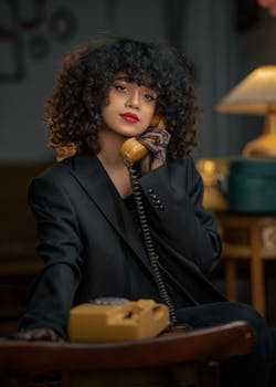 A stylish woman with curly hair talks on a vintage telephone in a warm, cozy setting.
