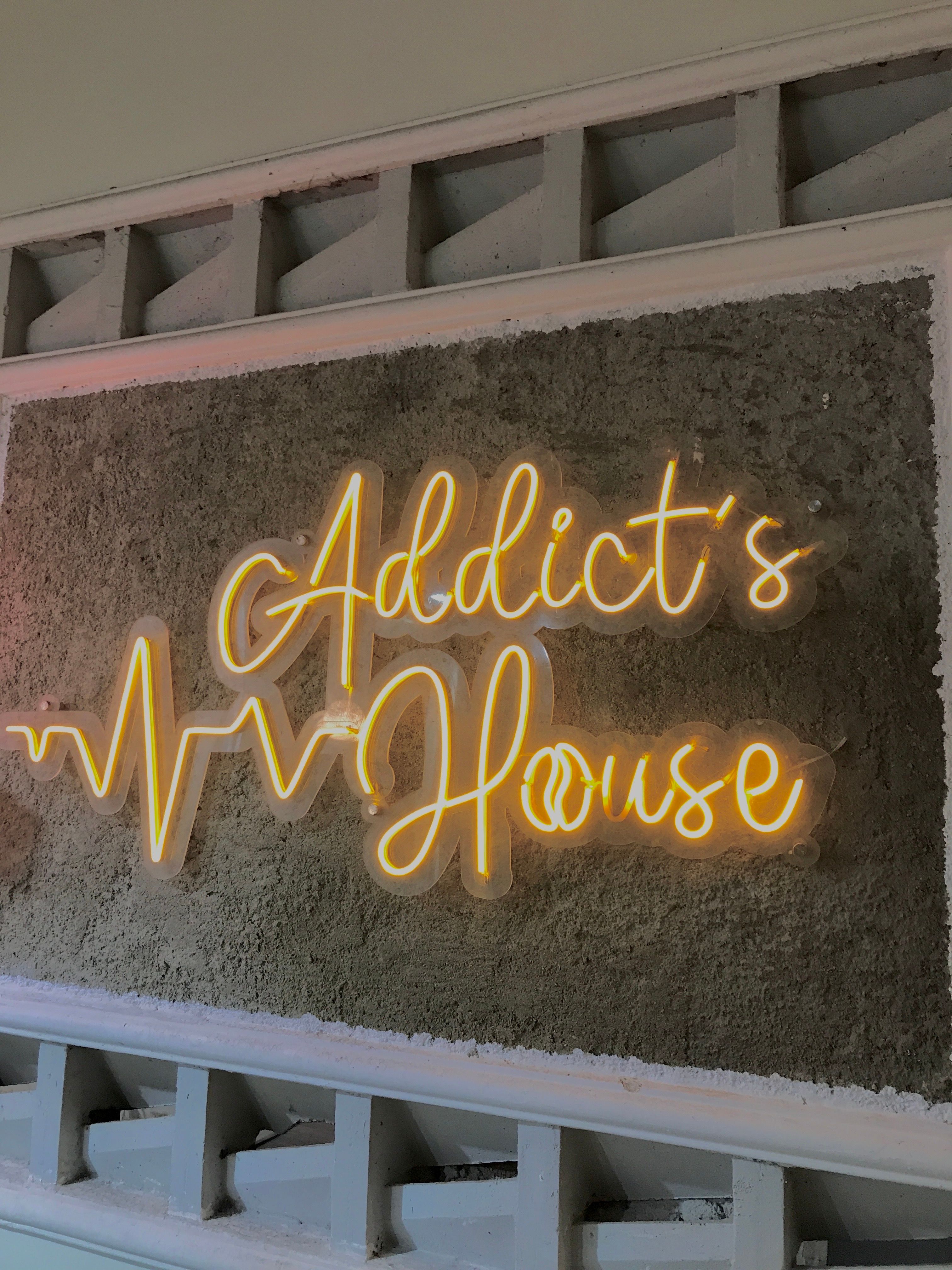 Illuminated neon sign 'Addict's House' with waveform design on concrete texture background.
