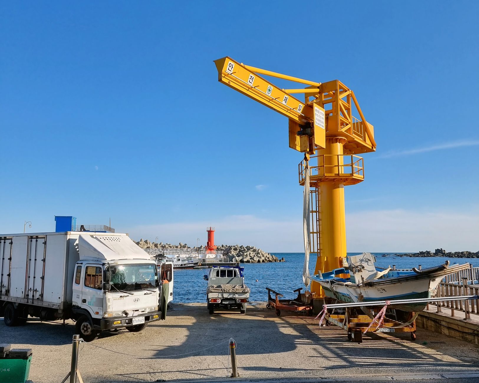 A vibrant harbor in Busan featuring a yellow crane, trucks, and a boat by the sea.