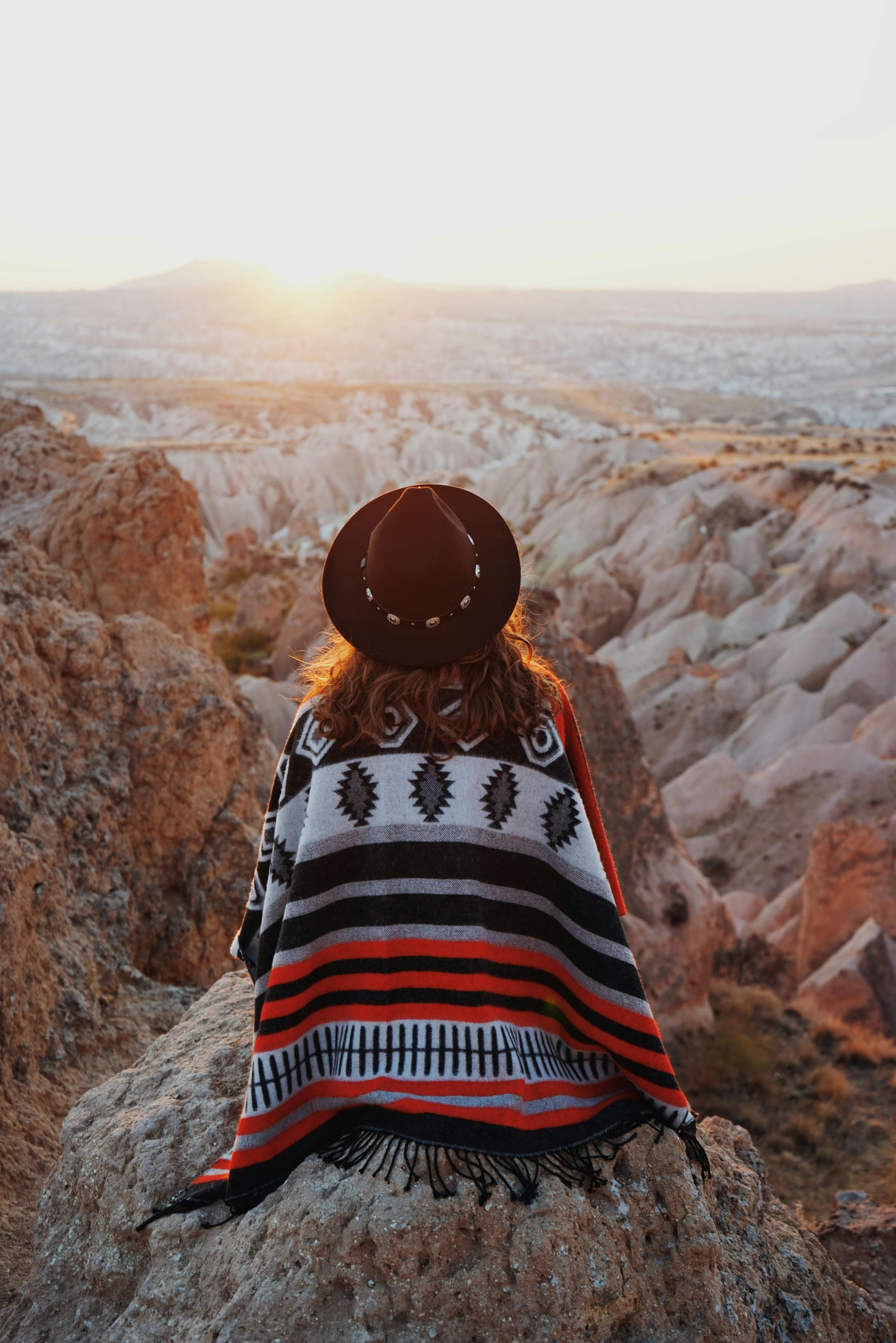 A woman in a hat and shawl enjoys a scenic sunset over the rock formations of Cappadocia, Turkey.