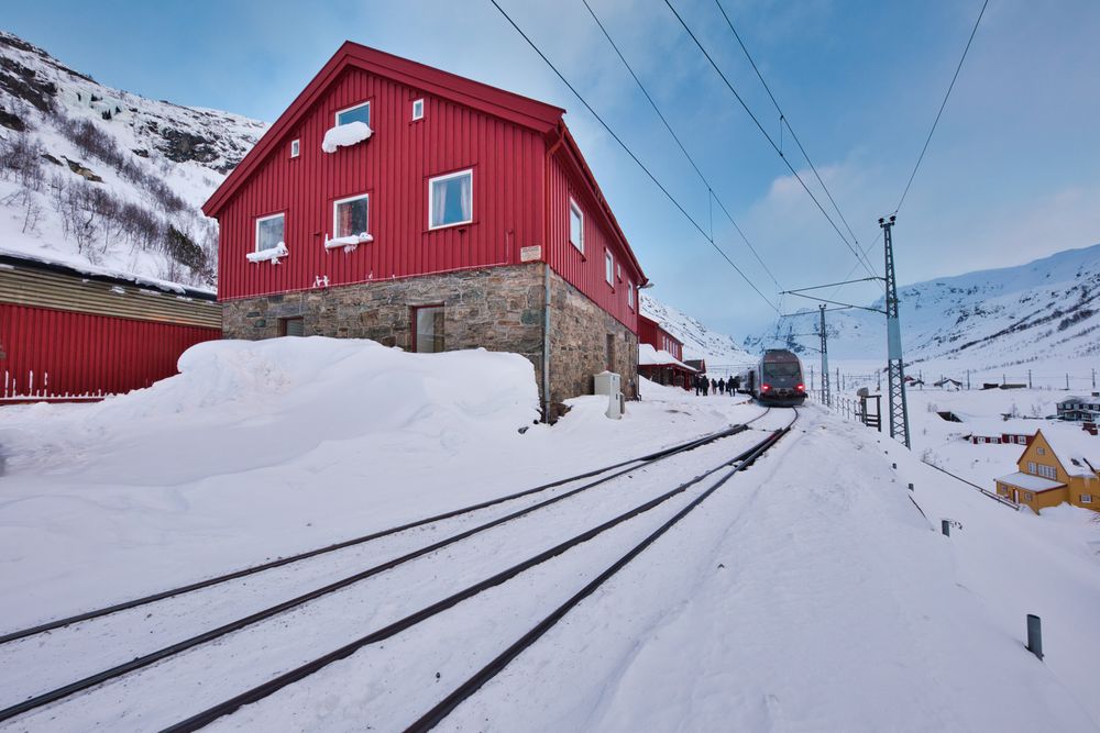 Casă roșie lângă șinele de tren, într-un peisaj de iarnă cu munți