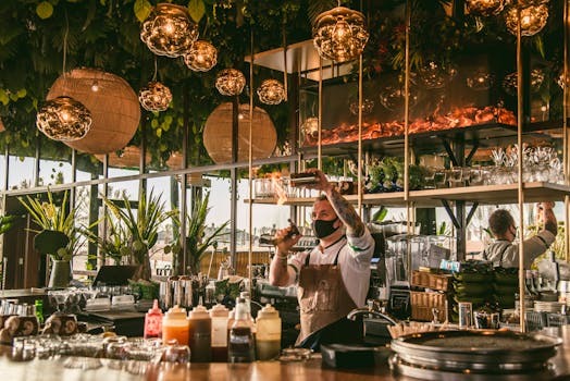 Bartender creates a flaming cocktail in a chic, plant-filled bar with modern decor and ambient lighting.