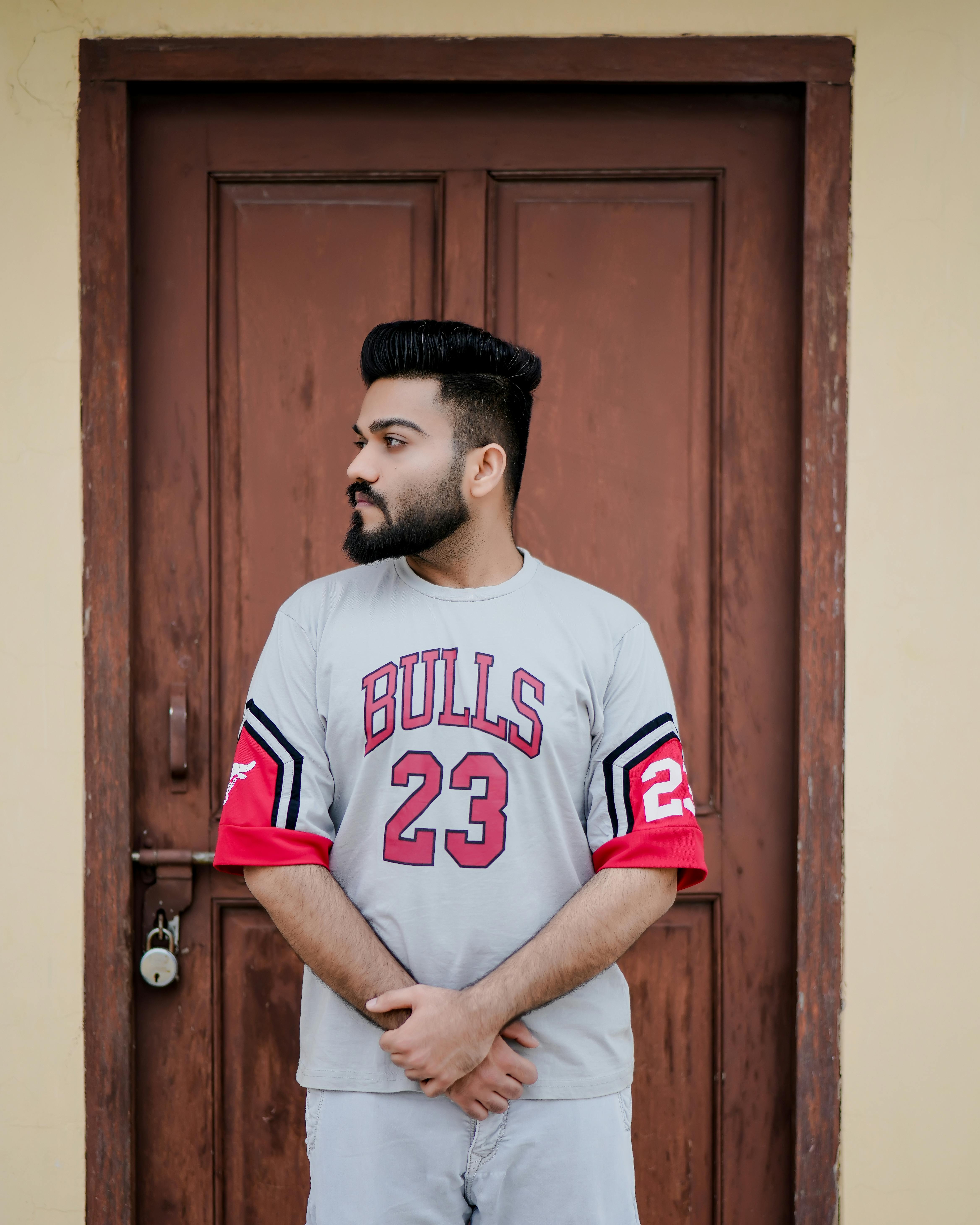 Young man with beard and mustache stands in front of a wooden door wearing a 'Bulls 23' shirt.