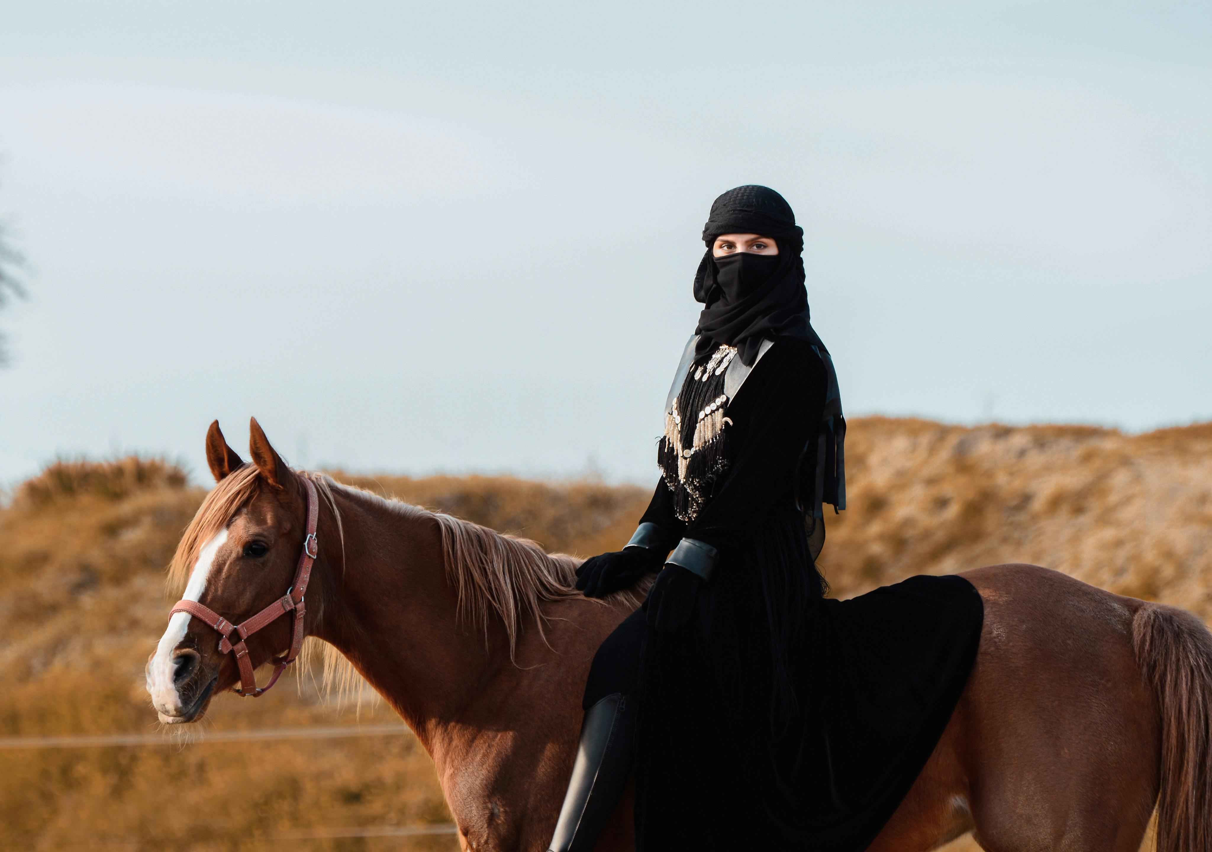 A Middle Eastern woman in traditional clothing riding a brown horse in a desert landscape.