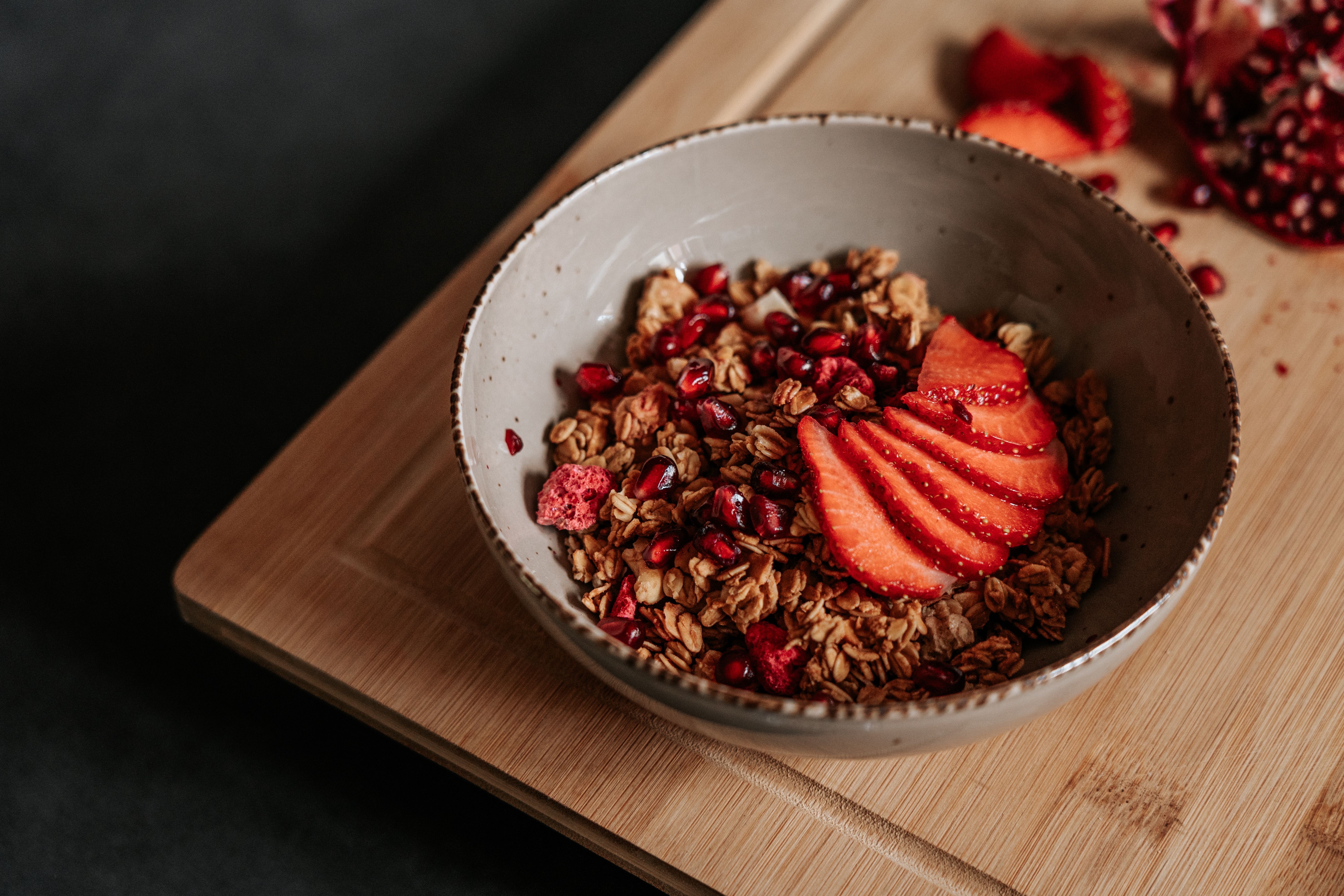 A close-up of a healthy breakfast bowl featuring granola, fresh strawberries, and pomegranate seeds on a wooden board.