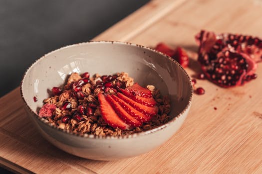 Delicious breakfast bowl with granola, strawberries, and pomegranate seeds on a wooden surface.