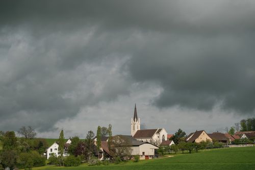 Free Idyllic rural village with historic church tower and dramatic skies, surrounded by lush green fields. Stock Photo