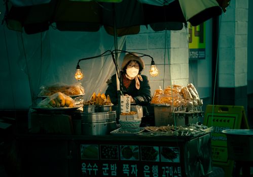 Asian woman vendor with mask selling food at night in Busan, South Korea.