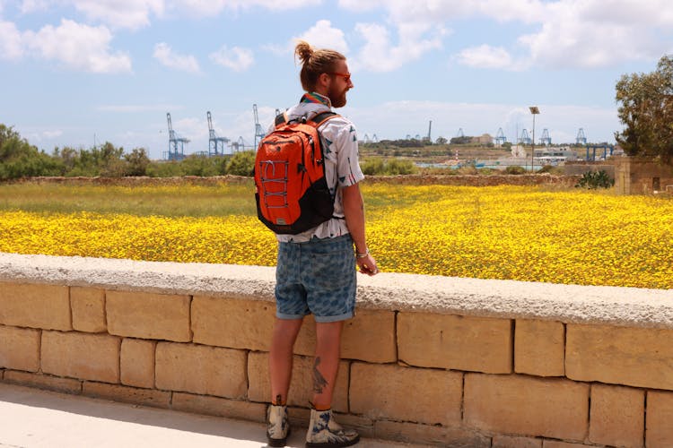 Back View Of Man With Backpack Standing By Meadow With Flowers