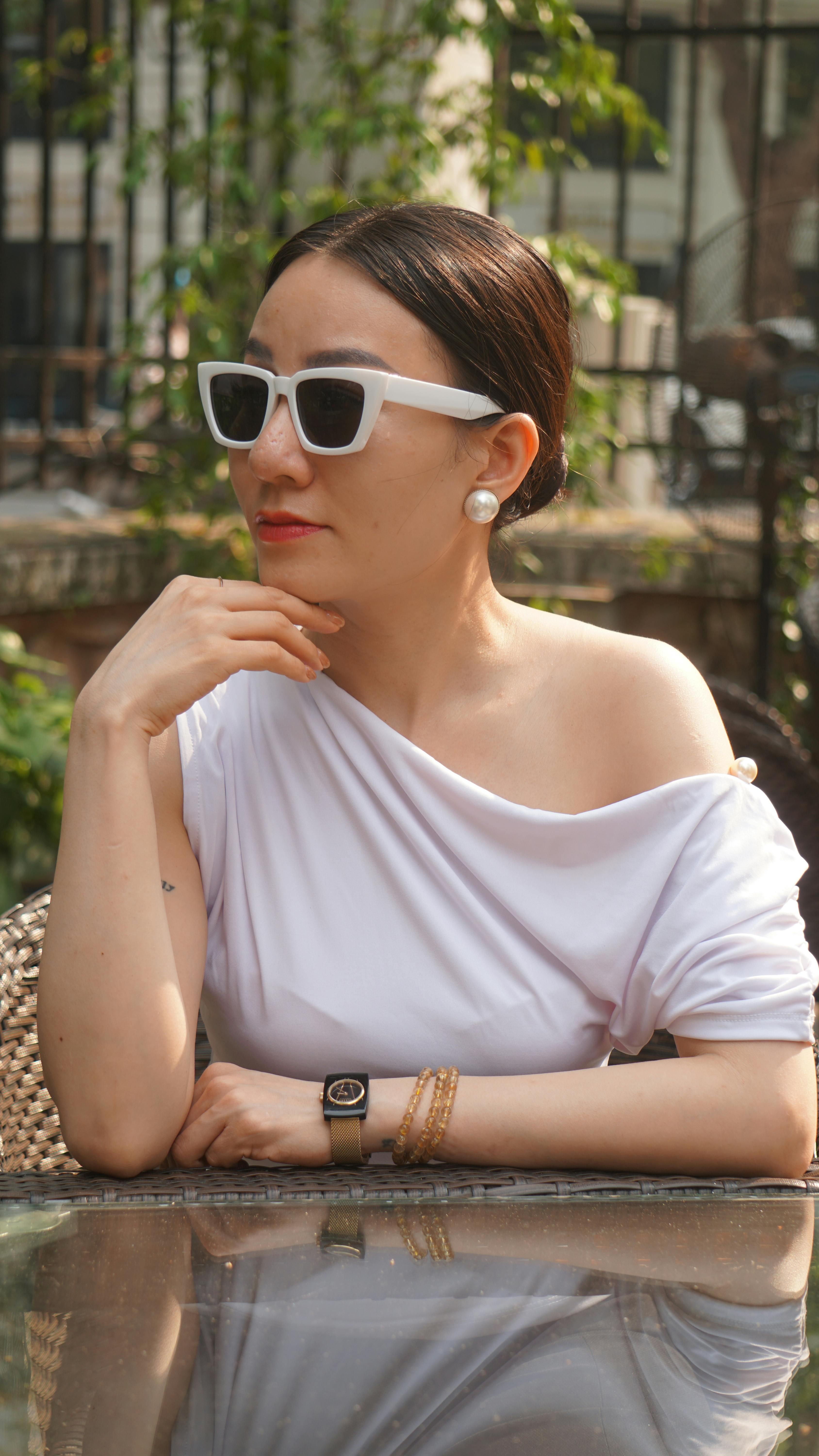 Stylish woman in white outfit and sunglasses relaxing at an outdoor cafe table on a sunny day.
