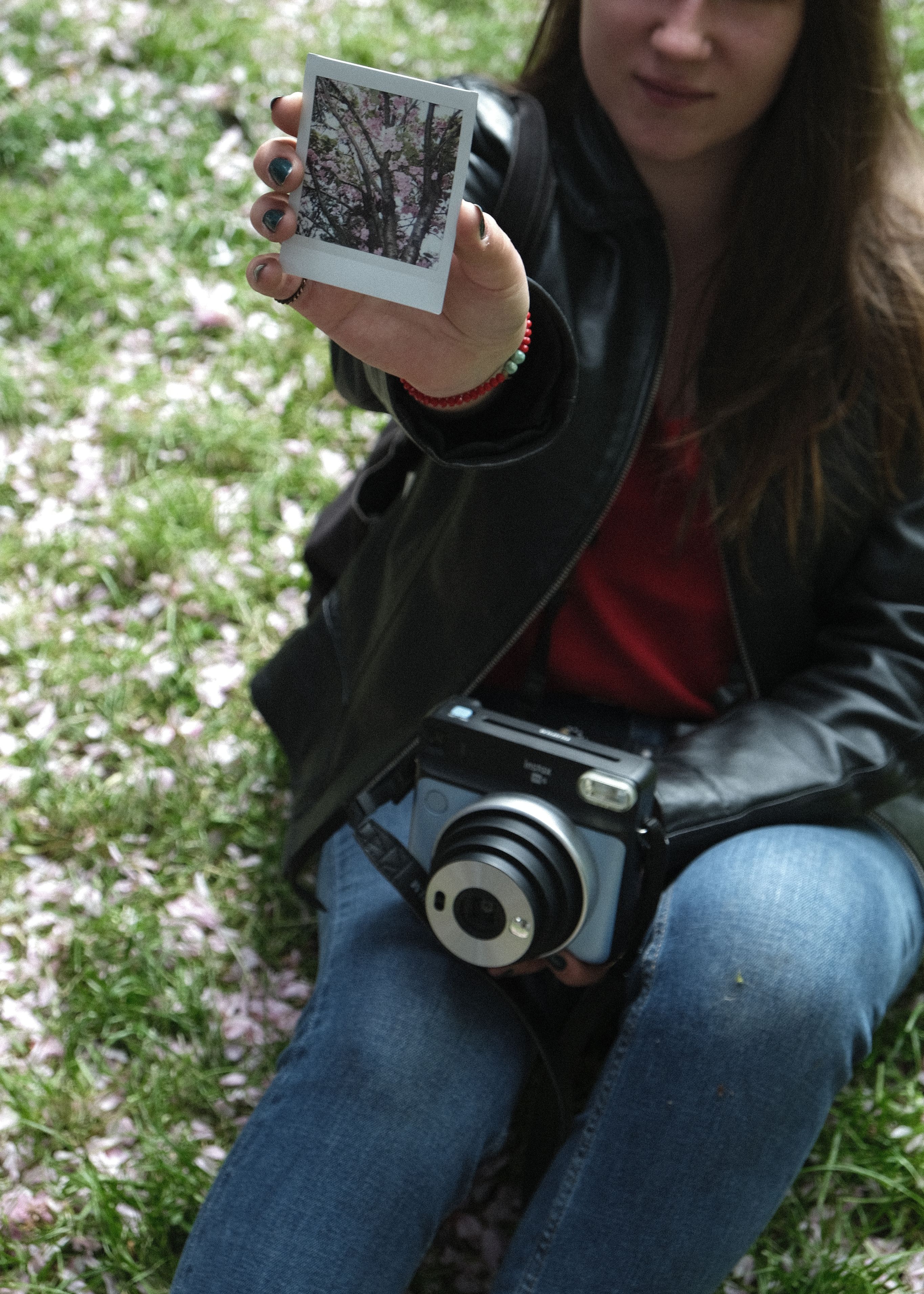 A woman with an instant camera sitting outdoors, showcasing a photo of blossoms.