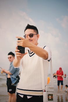 A young man in sunglasses taking a selfie at the beach on a sunny day.