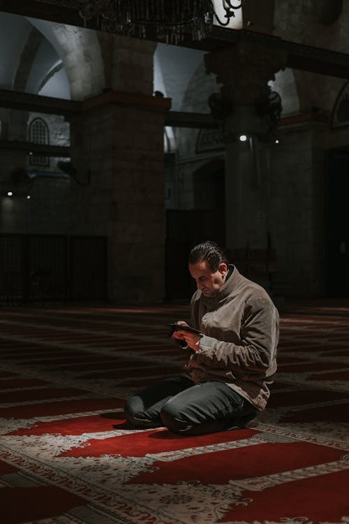 A man sitting on the floor in a mosque looking at his cell phone