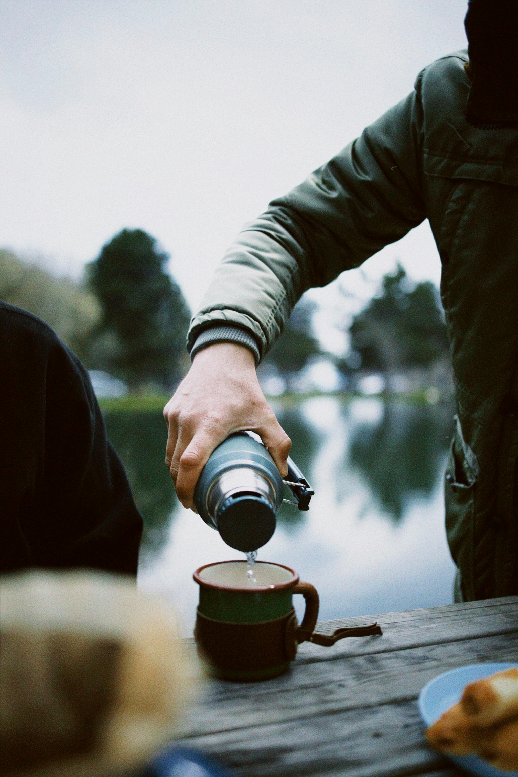 Person in jacket pours hot beverage into a mug by a scenic lake in Domaniç, Türkiye.