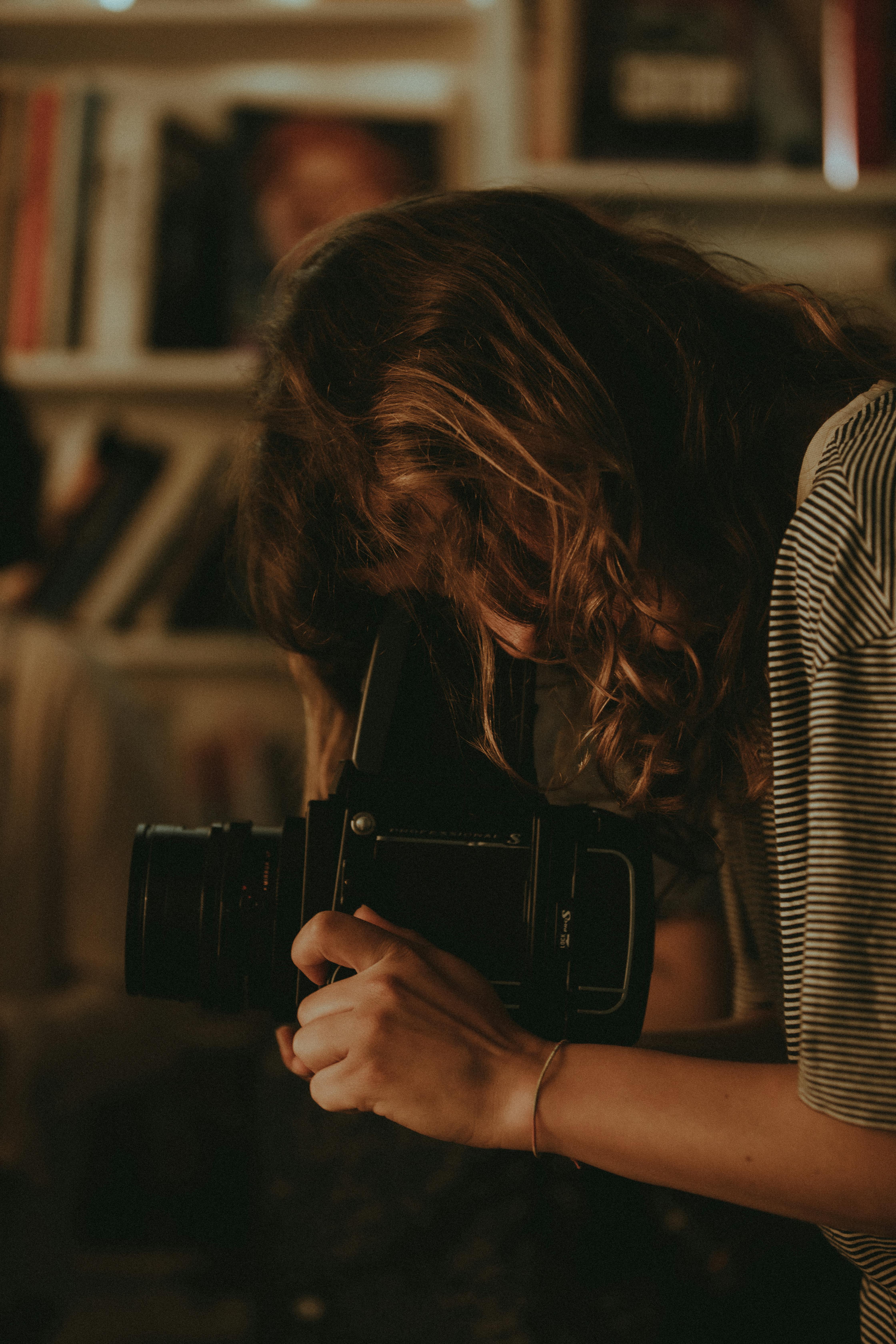 A woman with long hair takes a photo using an analog camera in a cozy indoor setting.
