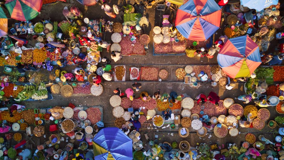 Colorful outdoor market bustling with people, fruits, and vegetables viewed from above.