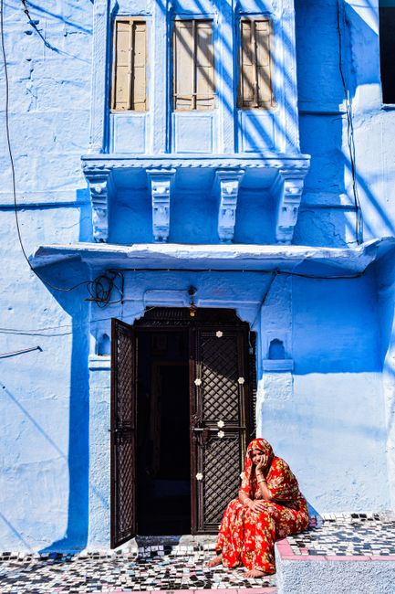 A vibrant depiction of urban life in Jodhpur, featuring a woman in traditional attire sitting by a striking blue house.