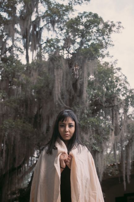 A woman standing outdoors wrapped in a blanket, with Spanish moss hanging from trees above.