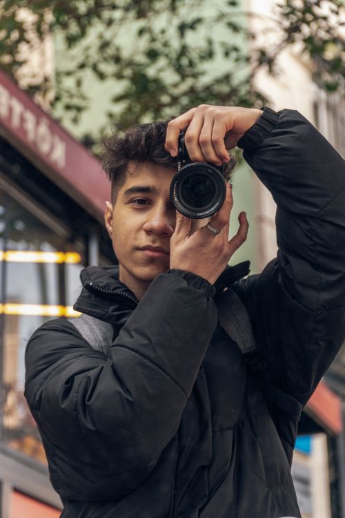 Free Young man in black jacket photographing on a bustling city sidewalk; urban exploration theme. Stock Photo