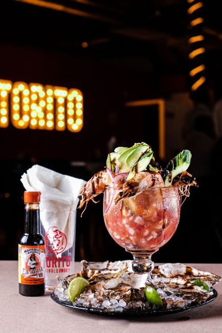 A glass bowl of pink shrimp ceviche de camarón mixed with diced tomato, cucumber, and cilantro, served with tostadas and lime wedges on the side.