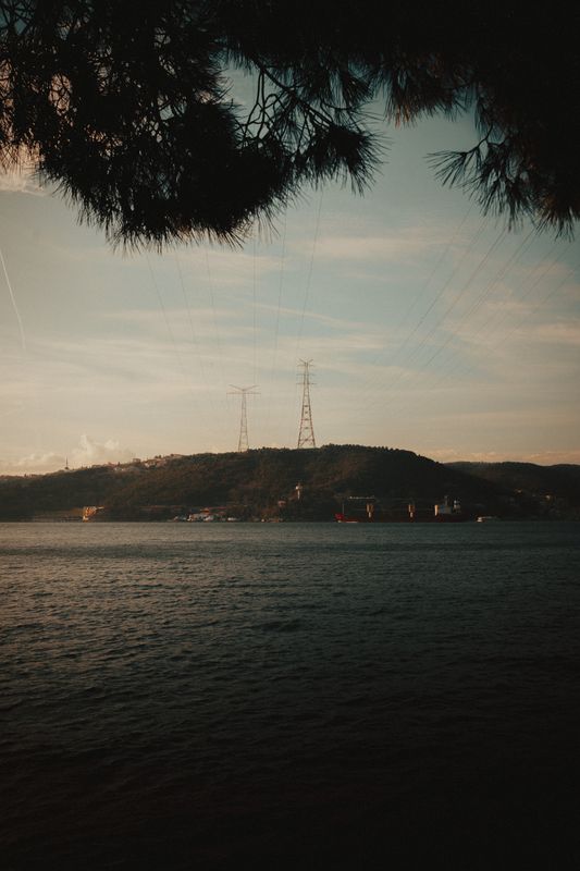 Alacakaranlıkta suyun karşısında elektrik direkleriyle tepelik bir kıyı