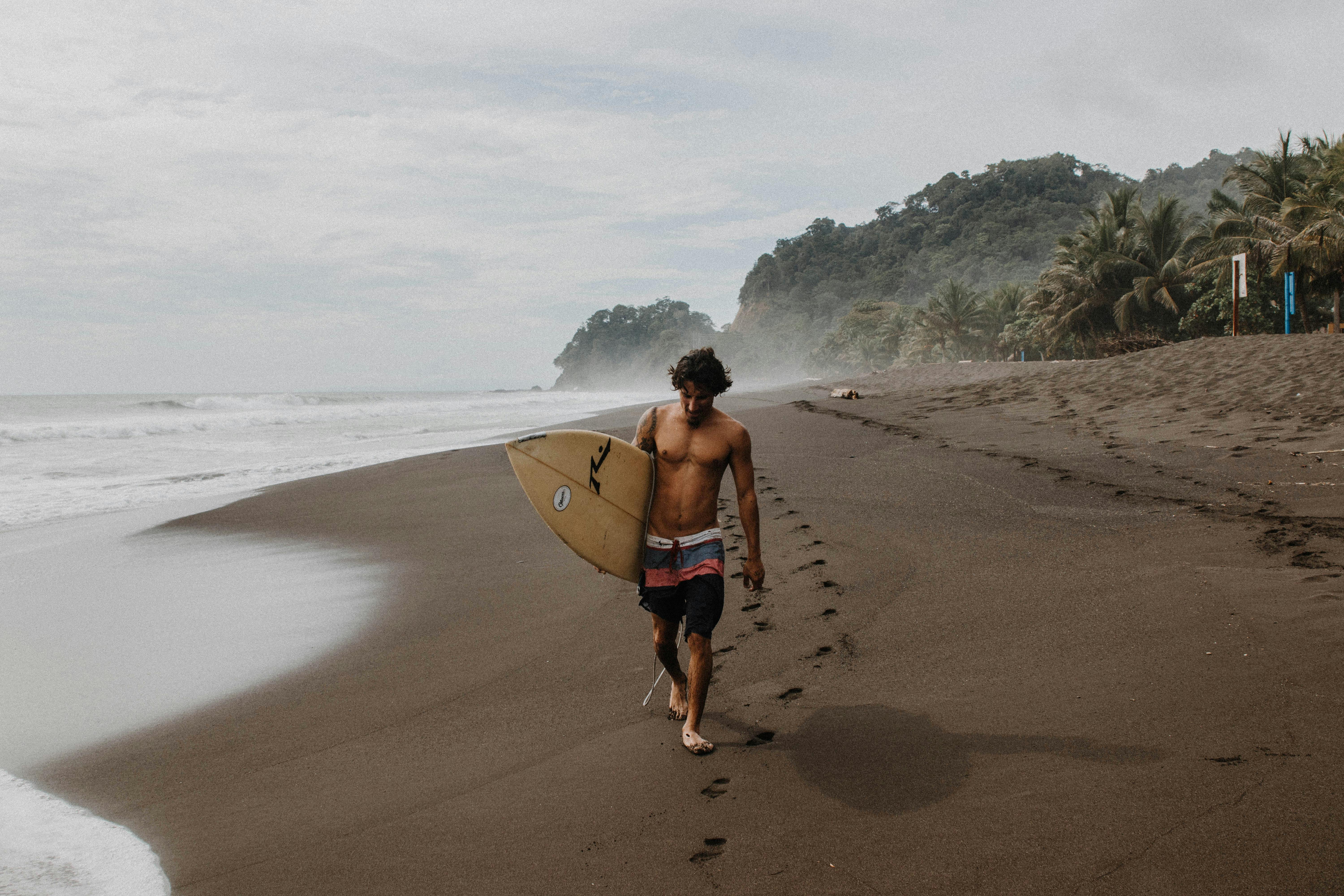 A surfer carrying a surfboard strolls along a tropical Costa Rican beach with palm trees and waves.
