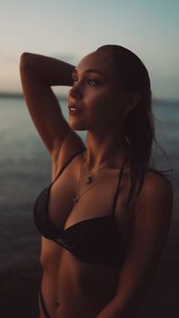 A woman in a bikini poses gracefully at the beach during twilight.