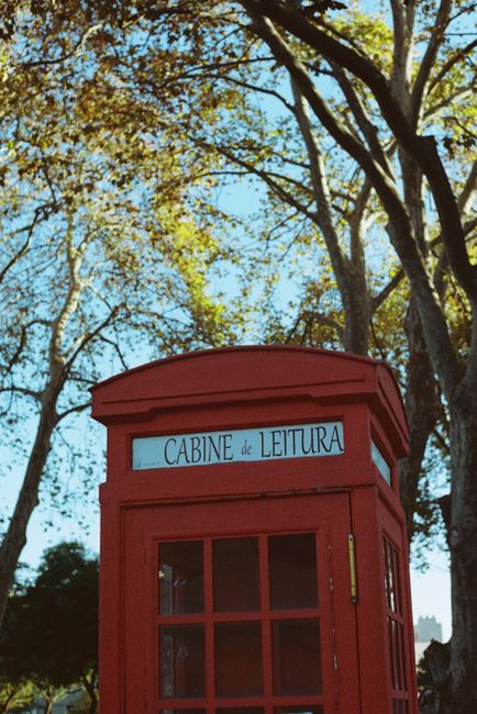 A vibrant red telephone booth amidst tall trees in Lisbon's urban park area.