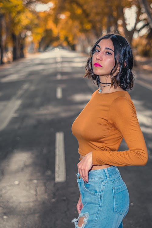 Young Woman Wearing Ripped Jeans Standing in the Middle of the Road between Autumnal Trees 