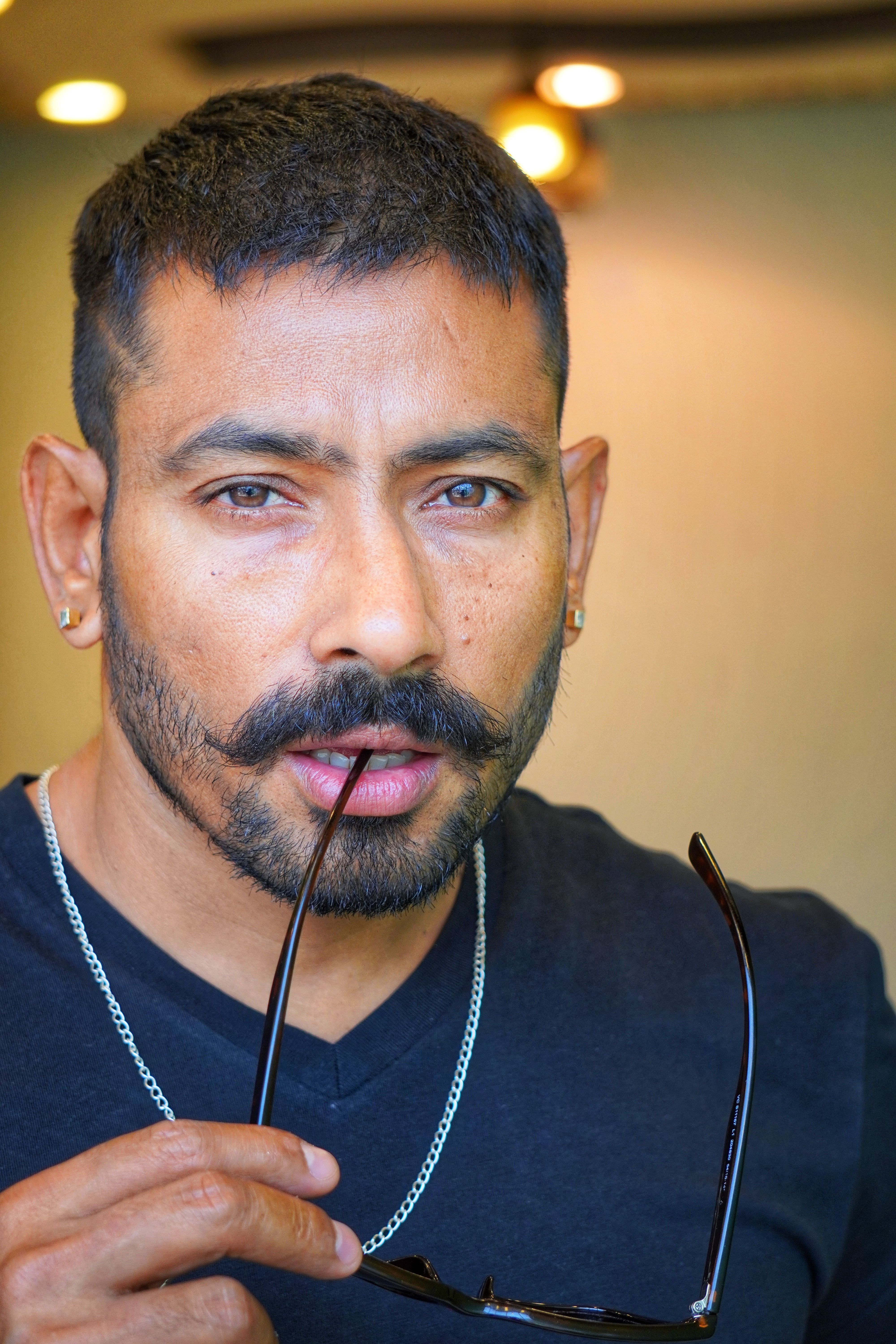 A close-up portrait of a stylish man with a beard and mustache holding sunglasses indoors.
