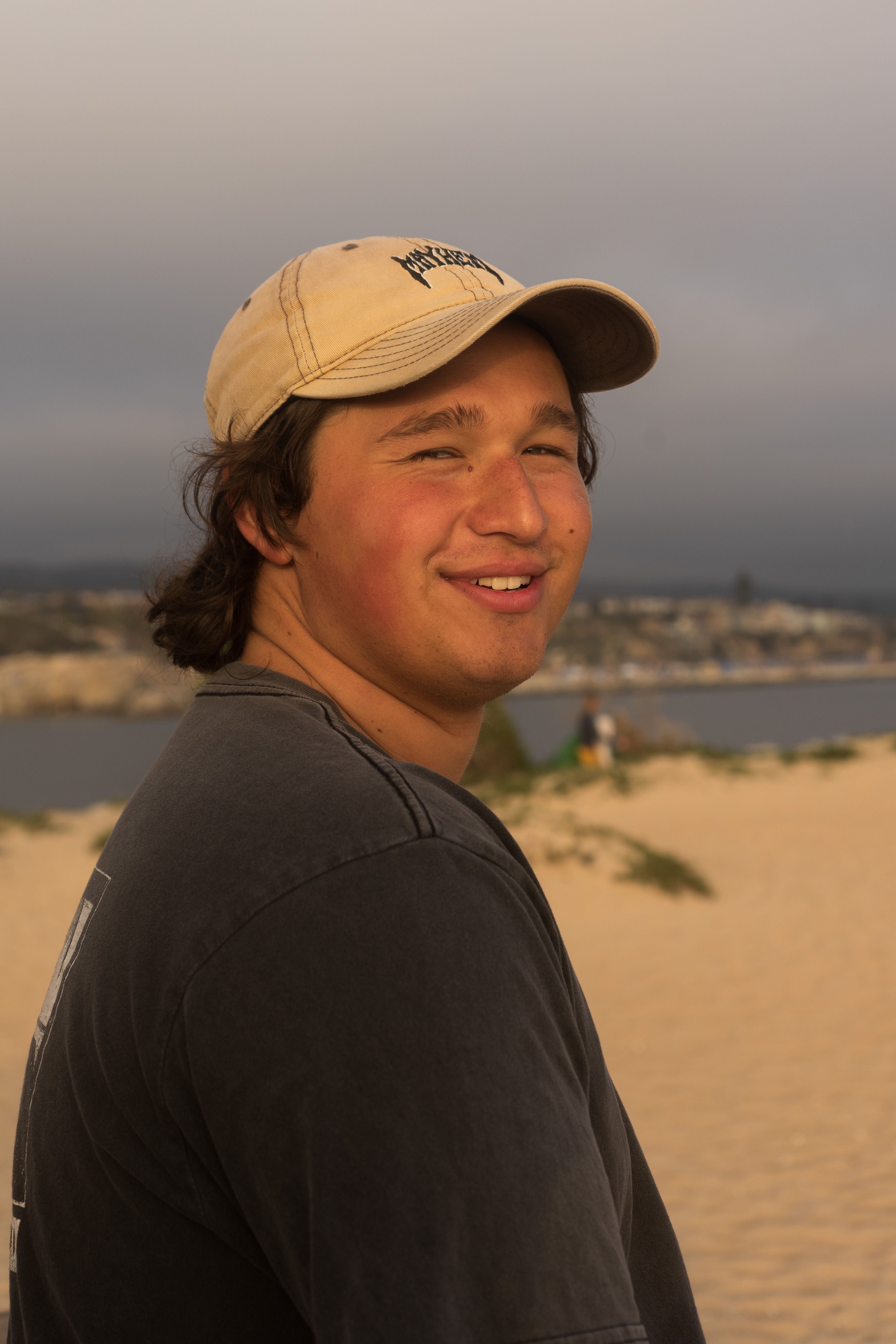 A cheerful young man wearing a cap smiles while standing on a sandy beach at sunset.