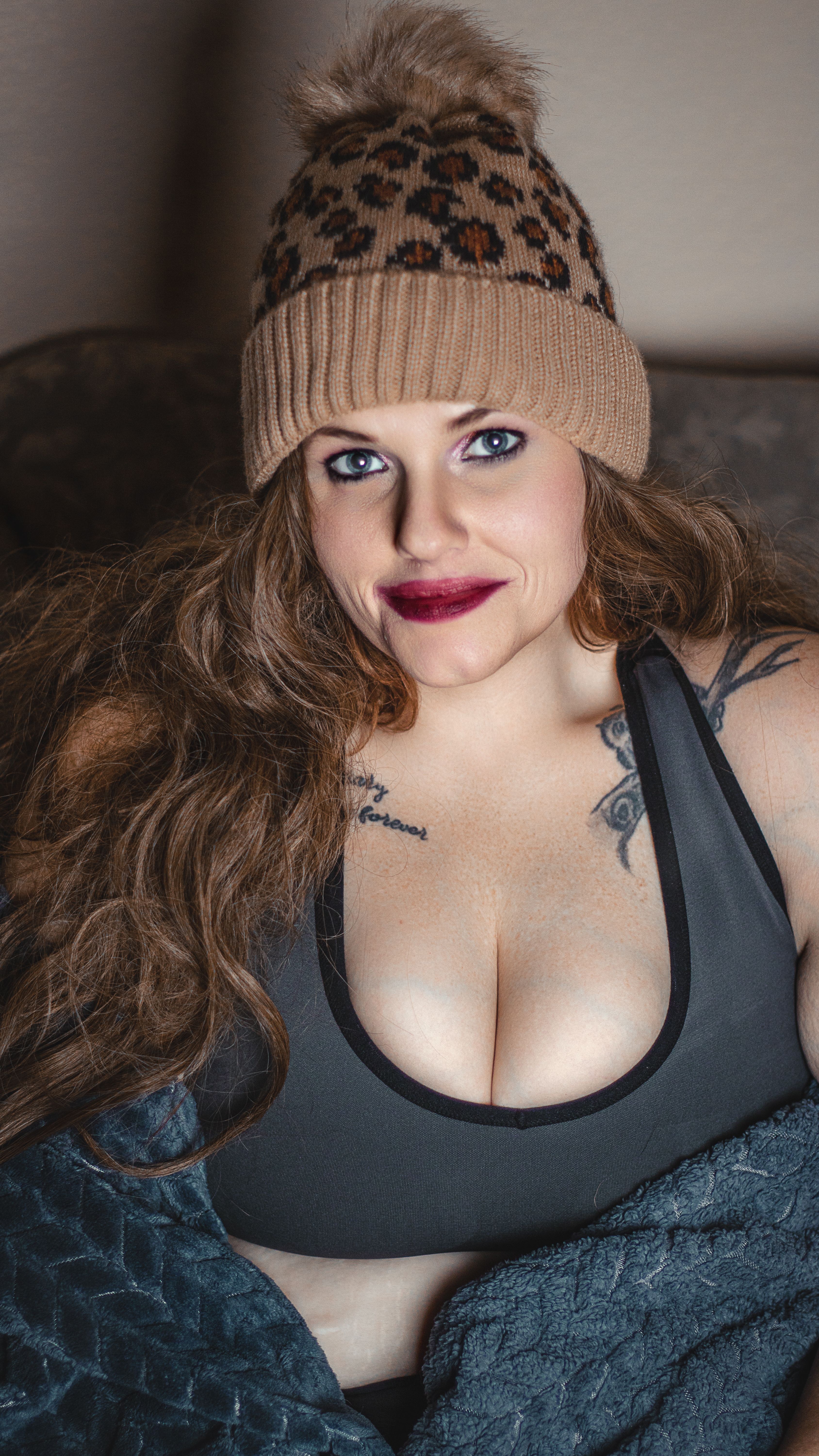 Portrait of a young woman with tattoos wearing a leopard print beanie and sports bra, smiling warmly.