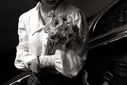Graceful woman in white blouse with bouquet of flowers stands on antique staircase, captured in black and white.