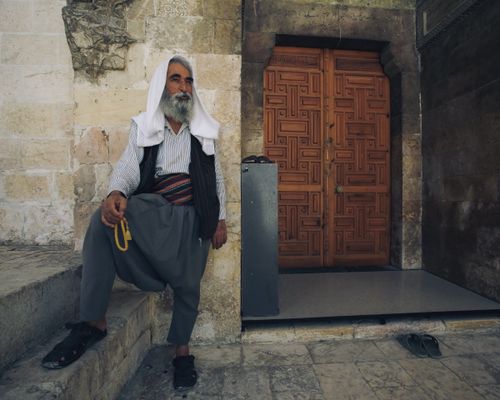 Free An elderly man in traditional attire rests near an ornate wooden door, showcasing cultural heritage. Stock Photo