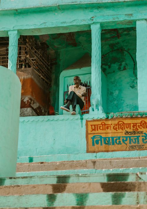 Free Elderly man sitting on turquoise steps in vibrant Varanasi street scene. Stock Photo