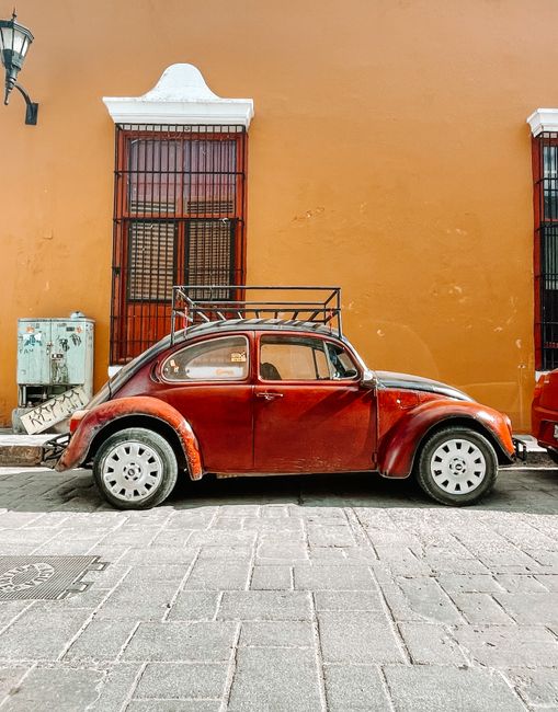 A classic red vintage car parked against a vibrant orange wall on a historic cobblestone street.