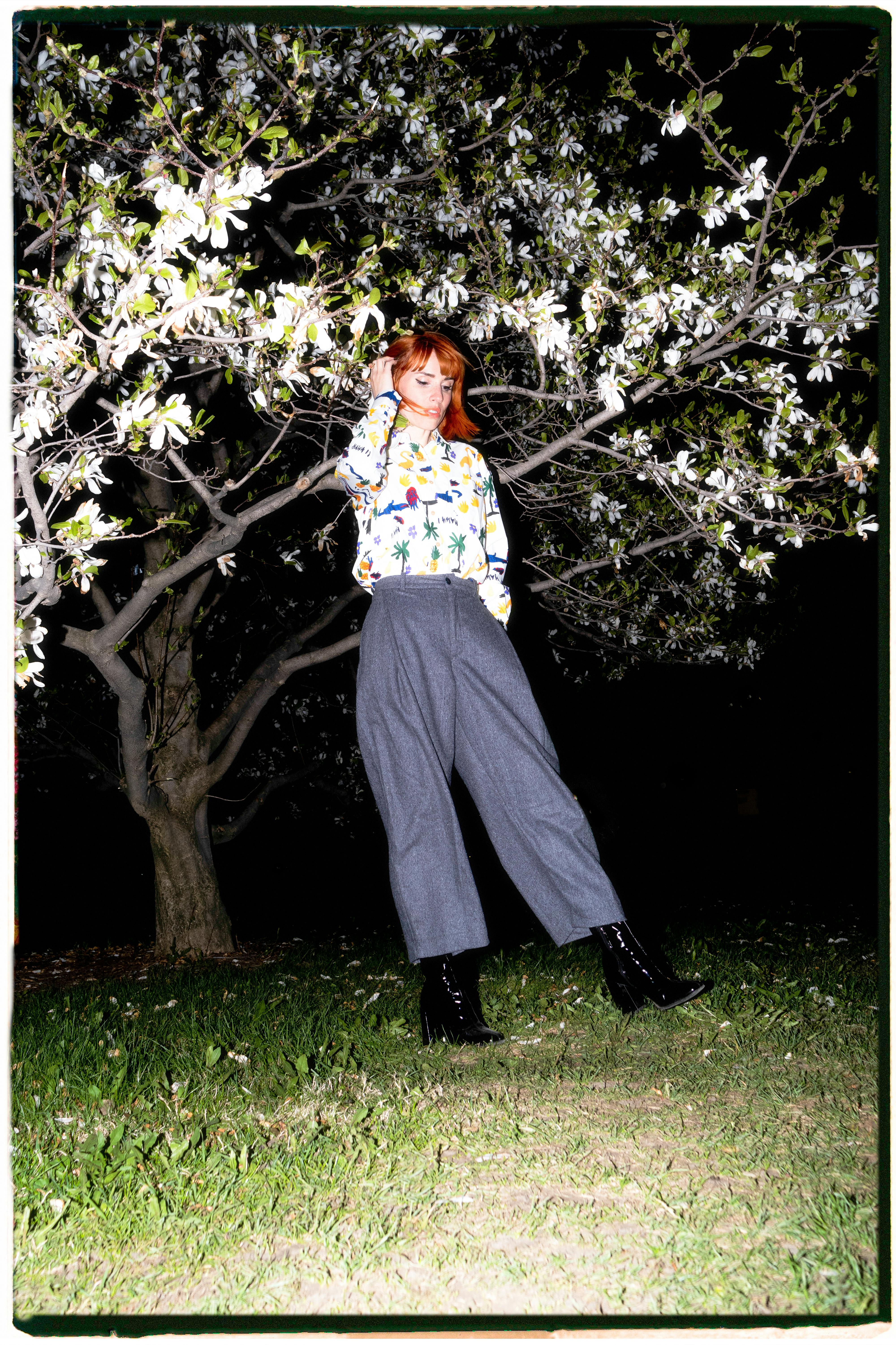 Chic woman with red hair in a floral blouse poses under a tree at night.
