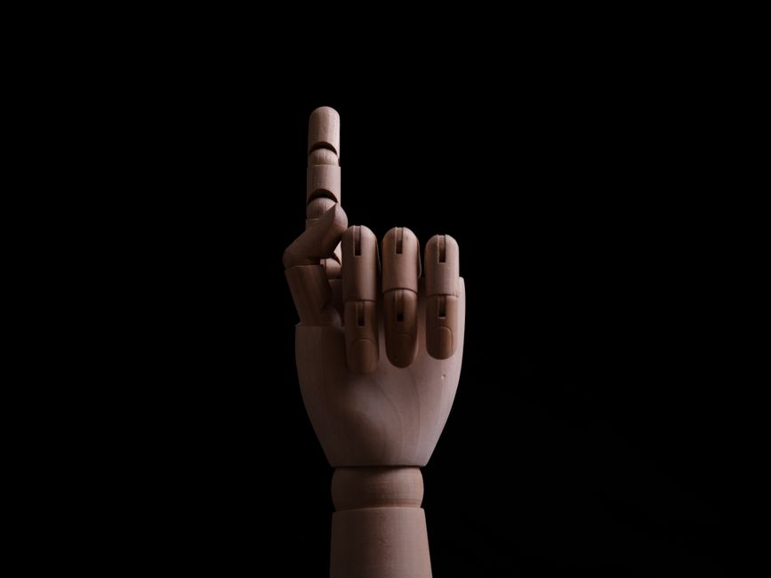 A close-up of a wooden articulated hand pointing upwards against a black background, conveying direction and expression.
