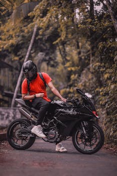 Biker in red shirt and helmet poses with a sleek black motorcycle on a scenic path.