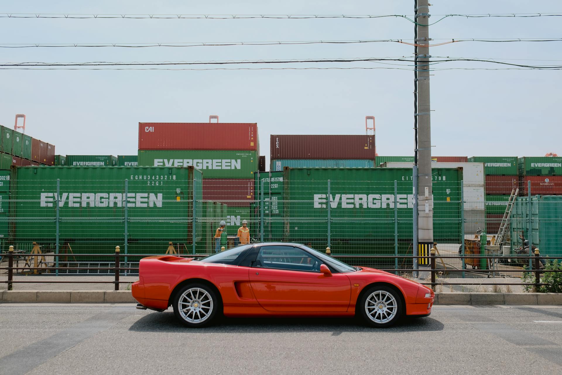 A vibrant red sports car parked near shipping containers labeled 'Evergreen' in a busy industrial area.