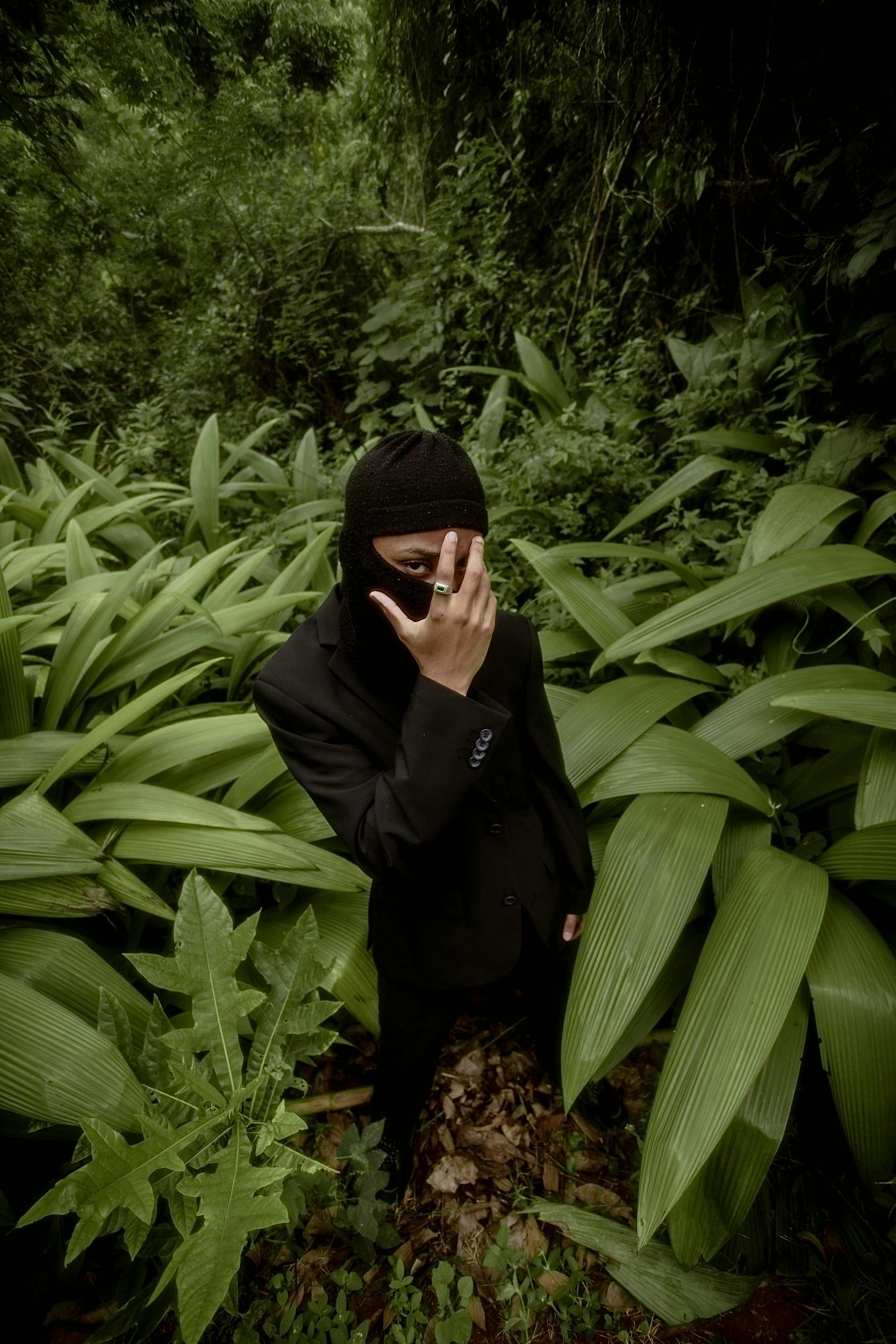 A figure in a black suit and balaclava poses amid lush greenery in a Brazilian jungle.