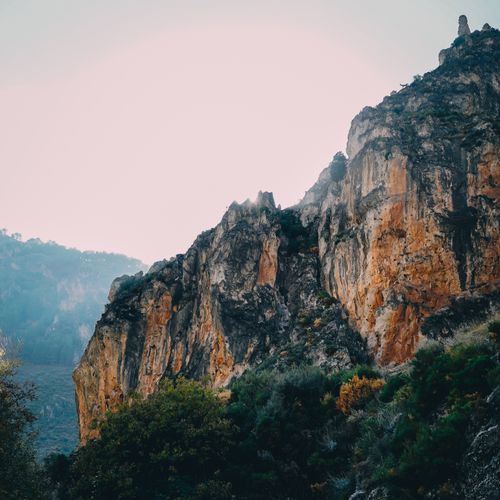 Free A breathtaking view of a rugged rocky cliff illuminated by the early morning sun, surrounded by lush greenery. Stock Photo