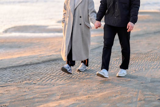 Romantic couple holding hands, walking on a beach during winter with sunlight.