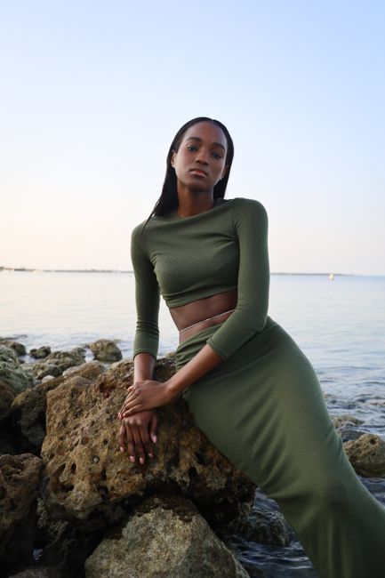 A woman in green sitting on rocks by the coast, exuding elegance and style.
